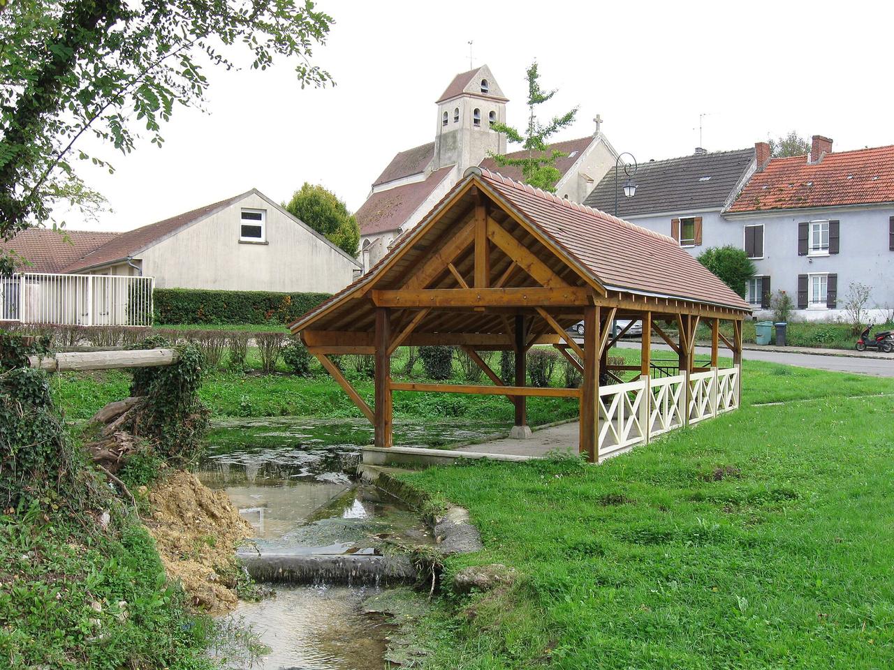 Lavoir en bois au bord d'un ruisseau avec église et maisons anciennes en arrière-plan.